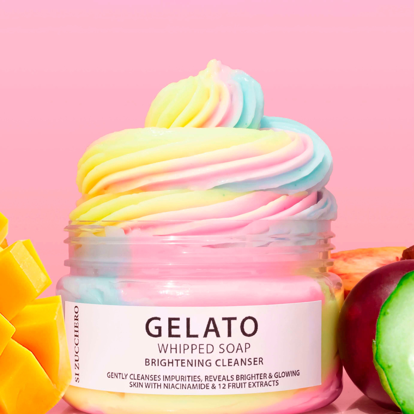 A jar of rainbow whipped soap cleanser beside mango and cucumber on pink